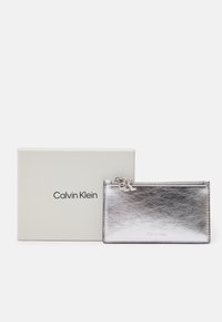 Sudraba metāla maks ar rāvējslēdzēju, ar logo un gludu virsmu, novietots blakus gaišas krāsas kastītei ar uzrakstu "Calvin Klein".