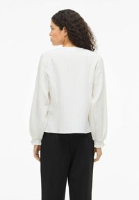 Hvid plisseret bluse med lange ærmer, indsnævrede manchetter og en afslappet pasform. Bærer over mørke bukser, set bagfra.