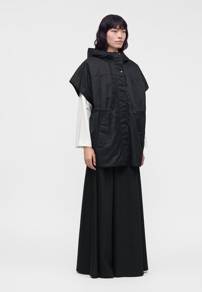 Femme portant un gilet sans manches à capuche noir sur un haut blanc à manches longues, associé à une longue jupe noire, debout devant un fond uni.