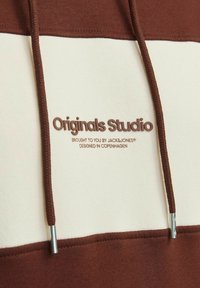 Brązowa i kremowa bluza z kapturem z drawstringiem i srebrnymi elementami. Na przodzie znajduje się wytłoczone logo "Originals Studio". Miękka tekstura materiału.