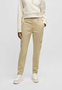 Pantaloni cargo beige con tasche laterali, realizzati in tessuto leggero, con una vestibilità dritta. Abbinati a sneaker bianche e a un maglione color crema.