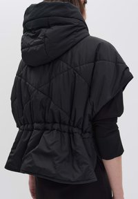 Gilet rembourré noir avec capuche, présentant un motif matelassé, une taille cintrée et des manches larges. Texture lisse avec une finition mate.