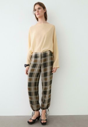 Femme debout portant une blouse beige à manches longues, un pantalon à carreaux marron et gris taille haute avec des poignets noués et des talons ouverts noirs.