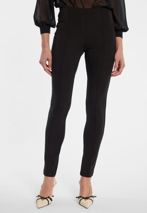Schwarze Slim-Fit-Hose mit vorderem Nahtdetail, getragen mit beigen spitzen Absätzen mit schwarzen Schleifen und dünnen Knöchelriemchen.