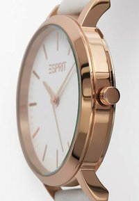 Roségoldene Uhr mit weißem Ziffernblatt, schlankem Profil, minimalistischem Design, hellfarbenem Armband und auffälliger Krone zur Zeiteinstellung.