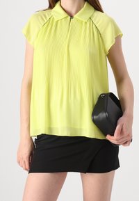 Blusa a pieghe gialla chiara con maniche corte e colletto, abbinata a una gonna nera e con una borsa nera.