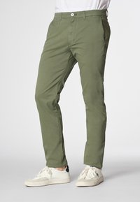 Modello che indossa pantaloni slim-fit verde oliva e sneakers bianche, posando su uno sfondo chiaro. Focus sulla parte inferiore dell'outfit.