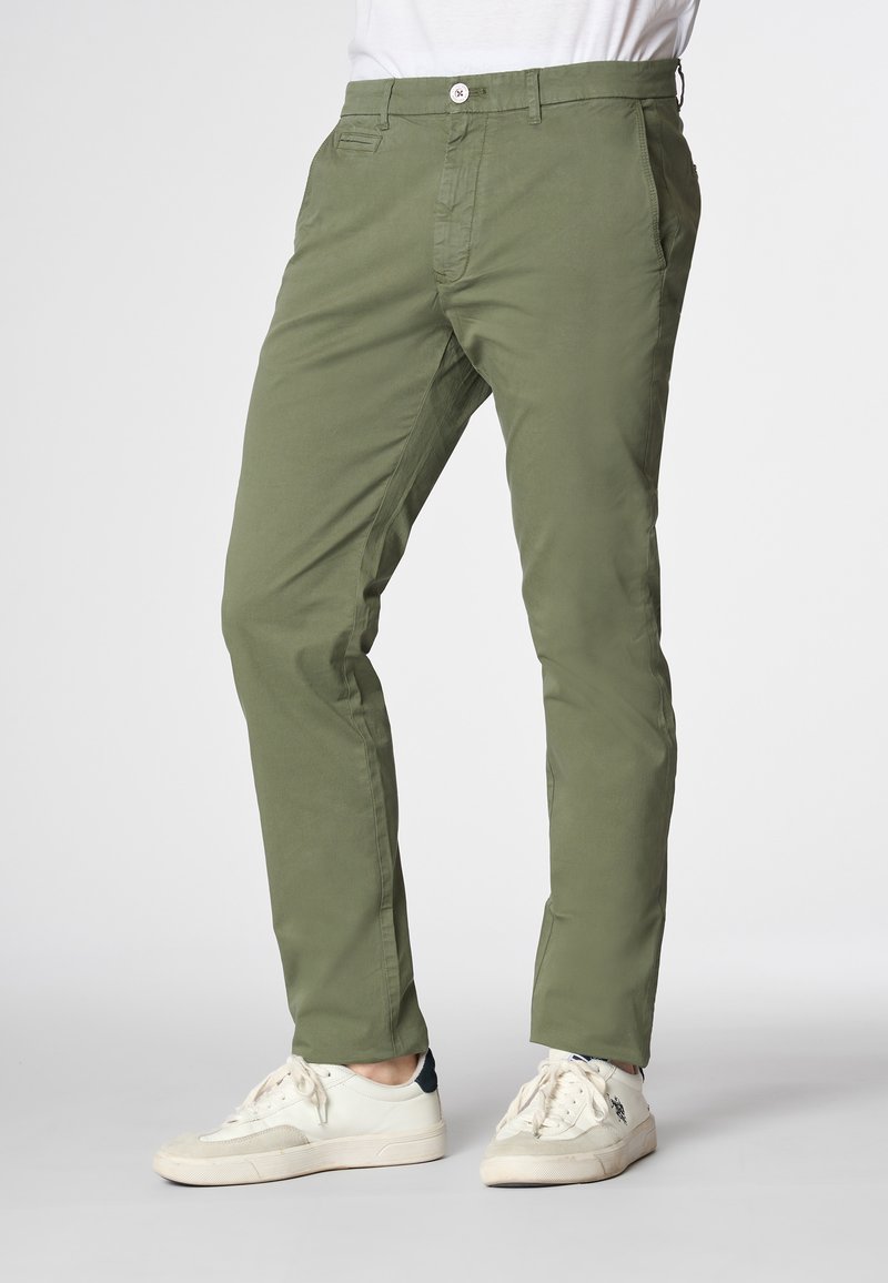 Modello che indossa pantaloni slim-fit verde oliva e sneakers bianche, posando su uno sfondo chiaro. Focus sulla parte inferiore dell'outfit.