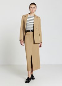 Femme portant un blazer beige et une jupe longue assortie avec une fente devant, un top rayé blanc et noir, une ceinture noire et des mocassins noirs.