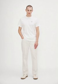 Bărbat în picioare, îmbrăcat cu un tricou alb cu un mic logo pe piept, pantaloni bej deschis și adidași albi, pe un fundal alb.