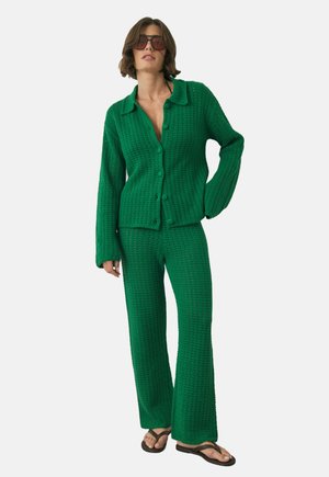 Vrouw in groene gehaakte knopen cardigan en bijpassende broek, met donkere zonnebril en zwarte flip-flops, staande met haar hand op haar heup.