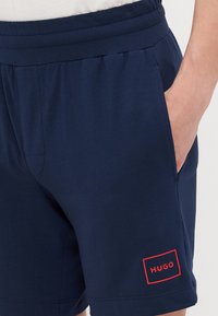 Marineblå shorts i et mykt stoff med elastisk midje, med et rødt rektangulært logo-lapp med teksten "HUGO" på venstre ben.