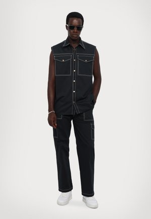 Zwarte mouwloze shirt met twee frontzakken, witte stiksels en gouden knopen, gecombineerd met bijpassende broek en witte sneakers.
