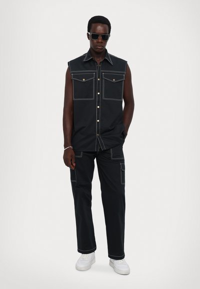 Zwarte mouwloze shirt met twee frontzakken, witte stiksels en gouden knopen, gecombineerd met bijpassende broek en witte sneakers.