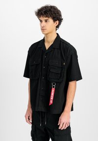 Zwart shirt met korte mouwen van katoen, met twee borstzakken, een knoopsluiting en een rode label die aan een zilveren ring hangt.