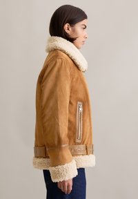 Tán kleurige schapenvacht jas met een crèmekleurige kraag en manchetten, voorzien van een zijzak met rits en een tailleband. Gladde textuur, oversized ontwerp.