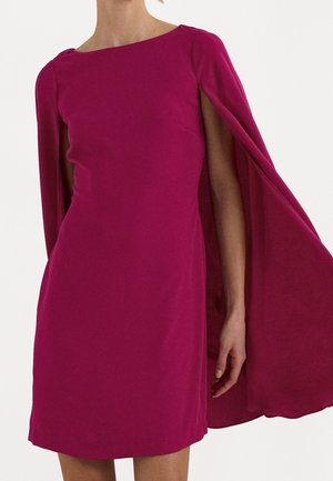 Femme portant une robe magenta sans manches avec des panneaux de cape attachés tombant sur ses bras, sur un fond uni.