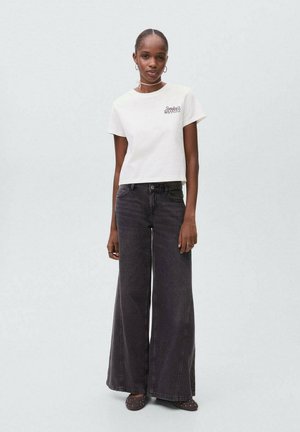 Femme portant un t-shirt court blanc avec le texte "Sunday’s Waffles", un jean noir ample, des boucles d'oreilles créoles et des chaussures brunes à clous, debout.