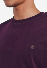 Violetti puuvillapaita, jossa on pyöreä kaula-aukko ja lyhyet hihat. Paidassa on rintataskussa pieni brodeerattu logo, joka on vastakkaisessa värissä.
