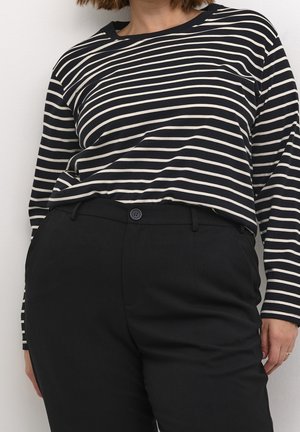 Persoon draagt een zwart-wit horizontaal gestreept shirt met lange mouwen, ingestopt in een hooggetailleerde zwarte broek met een knoopsluiting.