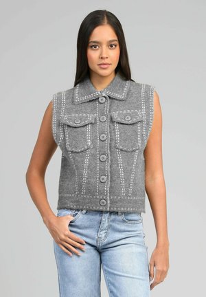 Jeune femme portant un gilet gris sans manches boutonné avec un motif cousu et un jean bleu clair, debout devant un fond uni.
