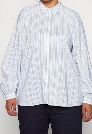 Kobieta w luźnej, białej koszuli z długim rękawem, z cienkimi niebieskimi pionowymi paskami i czarnych spodniach, stojąca ze zrelaksowanymi ramionami.