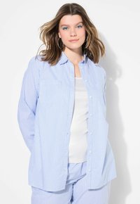 Camicia a righe blu chiaro e bianca con bottoni, maniche lunghe, taschino sul petto e vestibilità comoda. Indossata sopra un canottiere bianca.