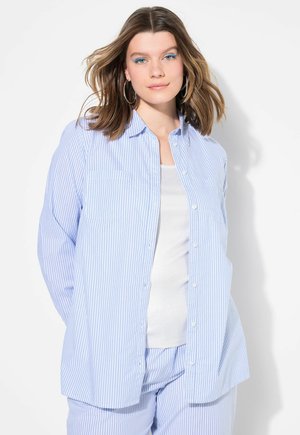 Chemise à rayures bleu clair et blanche, boutonnée, à manches longues, avec une poche poitrine unique et une coupe décontractée. Portée par-dessus un débardeur blanc.
