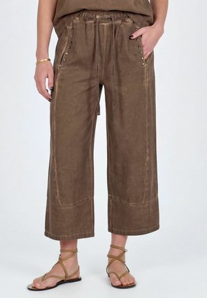 Pantalones marrones de pierna ancha y corte cropped con cintura con cordón y bolsillos con tachuelas, combinados con sandalias planas con tiras color cuero y una pulsera dorada.