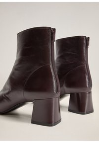 Par de botines de cuero marrón oscuro con tacones cuadrados y cremalleras traseras sobre un fondo neutro.