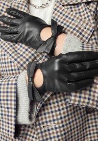 Schwarze Lederhandschuhe mit ausgeschnittenen Handgelenken und dekorativen Schleifen, getragen über einem karierten Mantel mit braunen und blauen Mustern. Weiche graue Fütterung sichtbar.