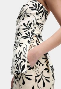 Witte stoffen top met zwart botanisch patroon, heeft een strapless ontwerp, zijsplitten en een handzak. Textuur, lichtgewicht materiaal.