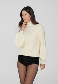 Suéter de punto crema con cuello alto y hombros caídos, combinado con pantalones cortos negros de lentejuelas. Textura suave, corte relajado y detalles de canalé.