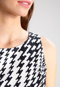 Läheltä otettu kuva naisen alahuulesta ja olkapäästä, joka on pukeutunut musta-valkoiseen houndstooth-kuvioiseen hihattomaan toppiin.