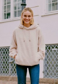 Světle béžová fleecová mikina s jemnou texturou, kapucí na šňůrku a předními kapsami, kombinována s modrými džíny. Volný a oversized střih.