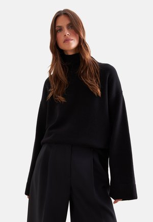 Femme aux longs cheveux bruns portant un pull à col roulé noir et un pantalon noir taille haute, regardant légèrement vers le haut sur un fond uni.