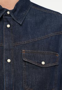 Ciemna koszula z denimu z ostrym kołnierzykiem, perłowymi zatrzaskami i dwoma kieszeniami na piersi. Kontrastujące pomarańczowe przeszycia podkreślają szwy.