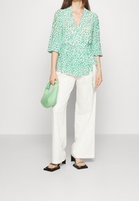 Grüne Leopardenmuster-Bluse mit einem Peplum-Silhouette, kombiniert mit weißen weit geschnittenen Hosen und schwarzen Quadratzehensandalen. Hält eine grüne Handtasche.