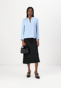 Blusa azzurra chiara a maniche lunghe con scollatura a V, abbinata a una gonna nera, con una piccola borsa nera in mano e indossando scarpe con tacco nere.