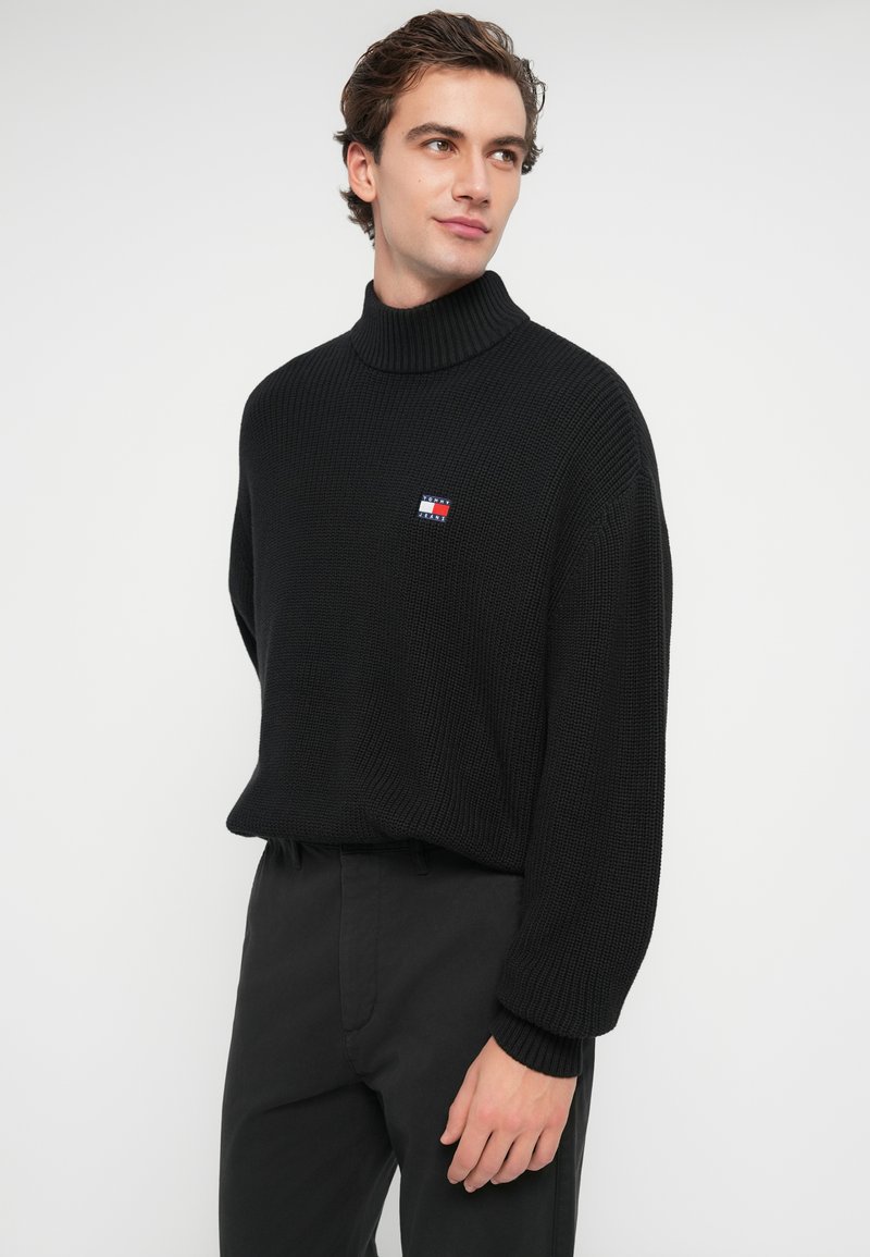 Zwarte gebreide turtleneck trui met een ribbelstructuur en een klein rood, wit en blauw logo op de borst. Gepaard met zwarte broek.