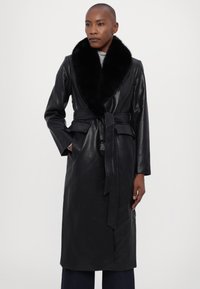 Μαύρο δερμάτινο παλτό με πλούσιο μαύρο γούνινο γιακά, ζώνη στη μέση, μακριά μανίκια και τσέπες; λεία υφή και ραμμένο σχέδιο.