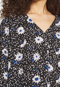 Černá květinová halenka s výstřihem do V, bílými sedmikráskami s modrými akcenty, knoflíkovým detailem a měkkou, texturovanou tkaninou.