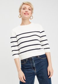 Maglione bianco con strisce orizzontali blu navy, scollo rotondo e maniche a tre quarti. Abbinato a jeans scuri a vita alta.