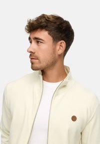 Joven con cabello corto castaño y barba recortada, llevando una chaqueta blanca con cremallera sobre una camiseta blanca, mirando hacia la izquierda contra un fondo liso.