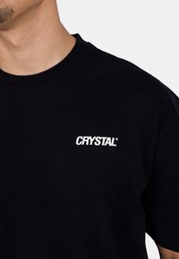 Černé bavlněné tričko s krátkým rukávem a kulatým výstřihem. Na přední straně je bílé logo "CRYSTAL" v moderním písmu.