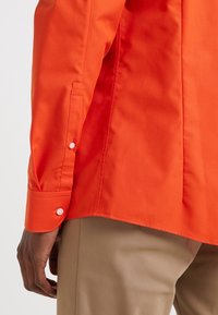 Persoon in een feloranje langemouwen shirt met geknoopte manchetten en beige broek, getoond vanaf de taille naar beneden op een witte achtergrond.