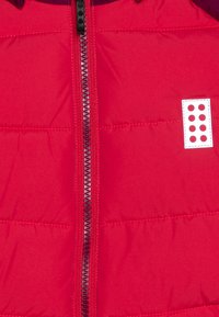 Czerwona kurtka puchowa z pełnym zamkiem błyskawicznym, pikowaną fakturą oraz małą białą łatą z czerwonymi kropkami po lewej stronie zamka.