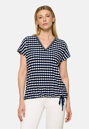 Femme portant un haut rayé bleu marine et blanc à col en V, manches courtes et ourlet à cordon de serrage, associé à un pantalon blanc.