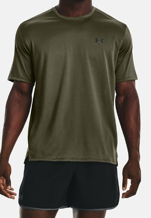 Athletischer Mann, der ein olivgrünes Kurzarm-Trainingsshirt und schwarze Sportshorts trägt und mit entspannten Armen steht.