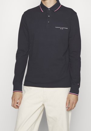 Mand iført en sort langærmet Tommy Hilfiger polo med hvide bukser, forfra, logo på brystet og stribede krave- og manchetkanter.